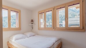 Un dormitorio con cama de madera, lámpara de pared y ventanas amplias con vistas a una montaña boscosa.