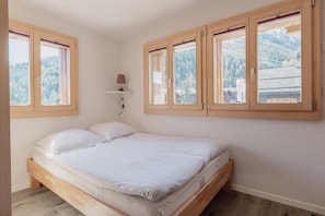 Ein Schlafzimmer mit einem hölzernen Bett, einer Wandleuchte und großen Fenstern mit Blick auf einen bewaldeten Berg.