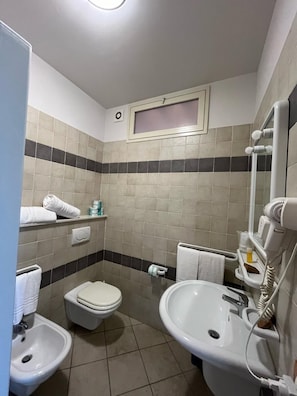 Une petite salle de bain comprenant des toilettes, un bidet et un lavabo. On y trouve également un porte-serviettes avec des serviettes, un miroir et un sèche-cheveux fixés au mur.