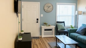 A living room with a wooden floor, a black side table, a green sofa, a white door, a clock on the wall, and a window with blinds.