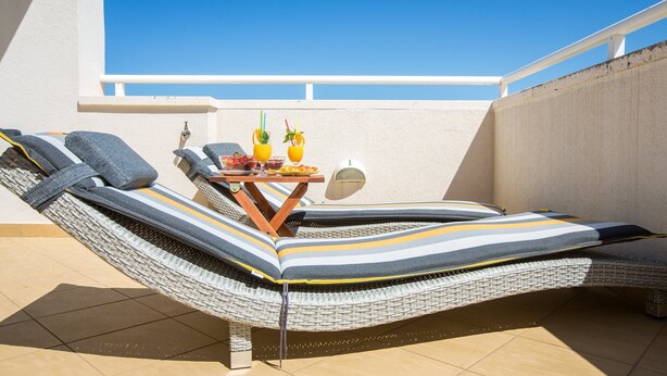 A sun lounger with striped cushions, a small table with drinks and a plate of food, and a white wall in the background. - Holiday Home Lapad Beach Royal (Dubrovnik)