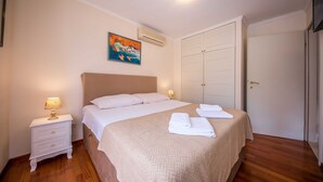 A neatly made bed with a beige blanket and pillows, a bedside table with a lamp, and a painting on the wall. - Holiday Home Lapad Beach Royal (Dubrovnik)