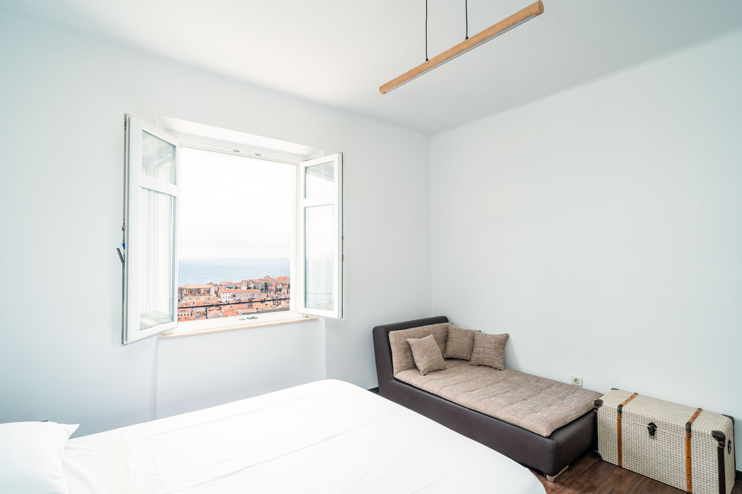 Une chambre à coucher avec un lit, une fenêtre dont le volet est ouvert, une vue sur des immeubles, un petit canapé et une valise.
