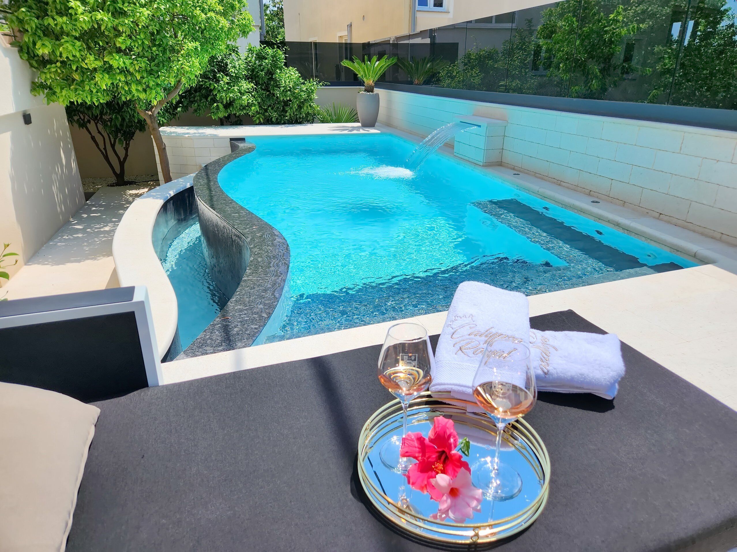 A pool with a water feature, a glass table with a tray, a glass of wine, and a towel with "Congratulations Royal" embroidered on it.