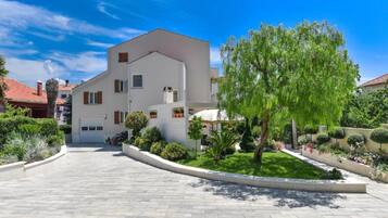 Una casa moderna de dos pisos con fachada blanca, entrada curva y un jardín cuidado con diversas plantas y un árbol grande.