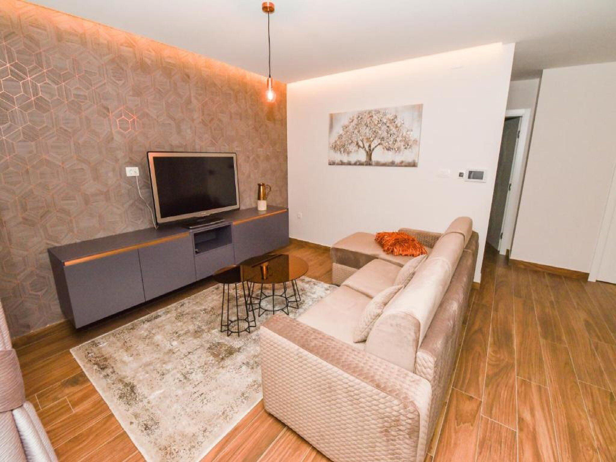 Uma sala de estar moderna com televisão de tela plana sobre um console cinza, sofá bege, tapete estampado e um quadro de árvore fixado na parede.