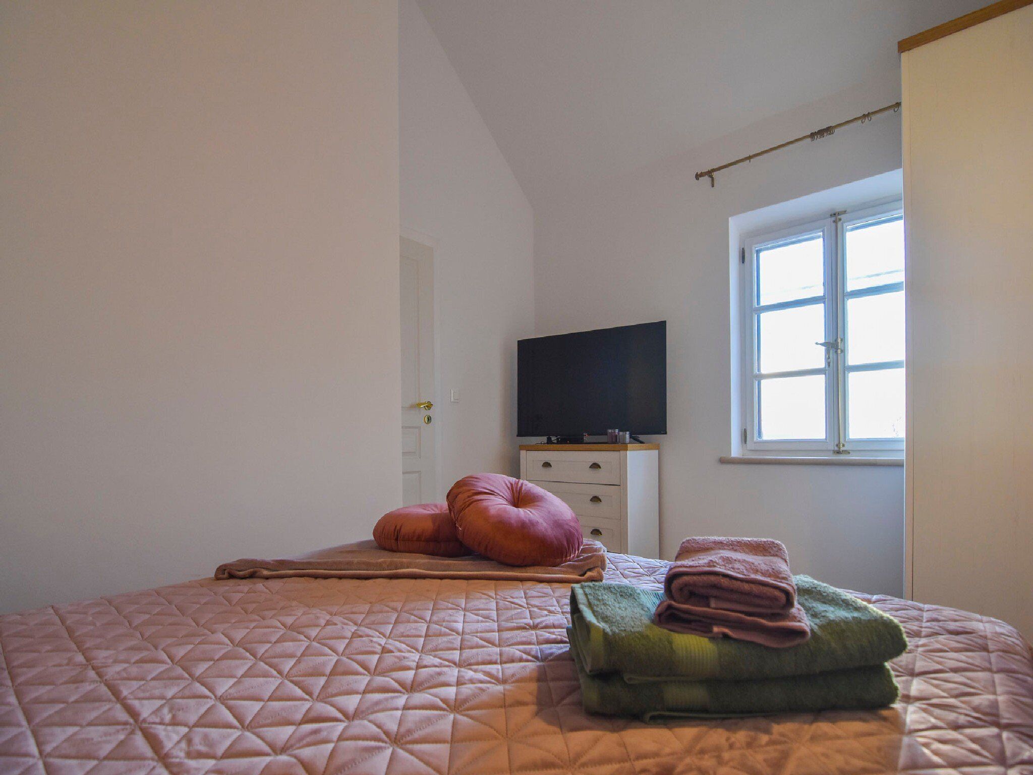 Une chambre à coucher avec un lit, des oreillers, un téléviseur posé sur une commode et une fenêtre.