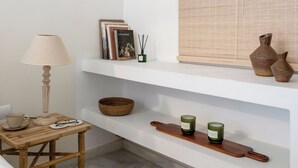 Habitación minimalista con una mesa de madera, una lámpara, una estantería con libros y adornos, y un ventanal con persianas.