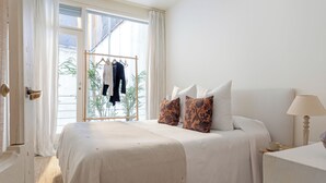 Un dormitorio con cama, almohadas, una mesita de noche, una lámpara y una ventana con cortinas.