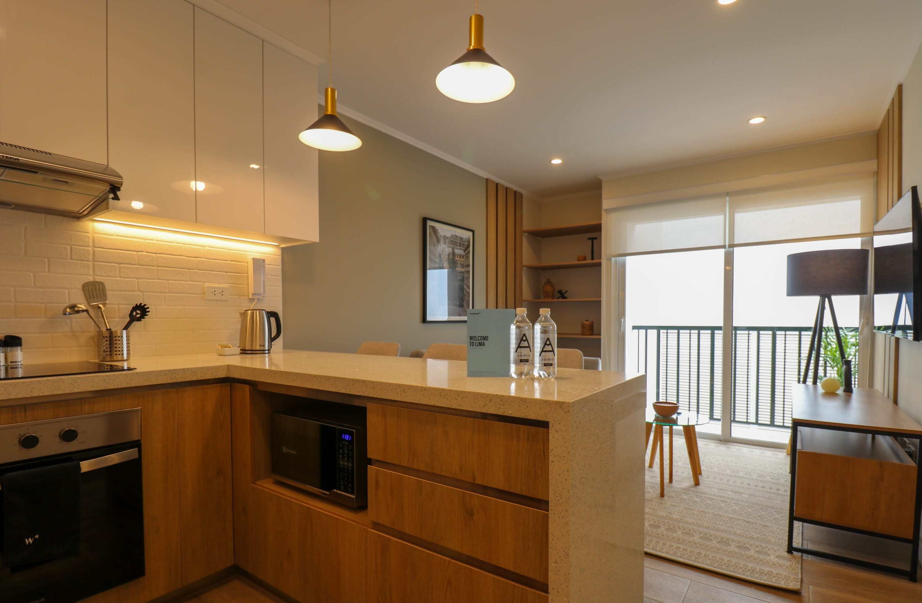 A modern kitchen with wooden cabinets, a built-in oven, and a coffee maker. There is a small island with a welcome sign and two bottles. The kitchen leads to a balcony with a view.