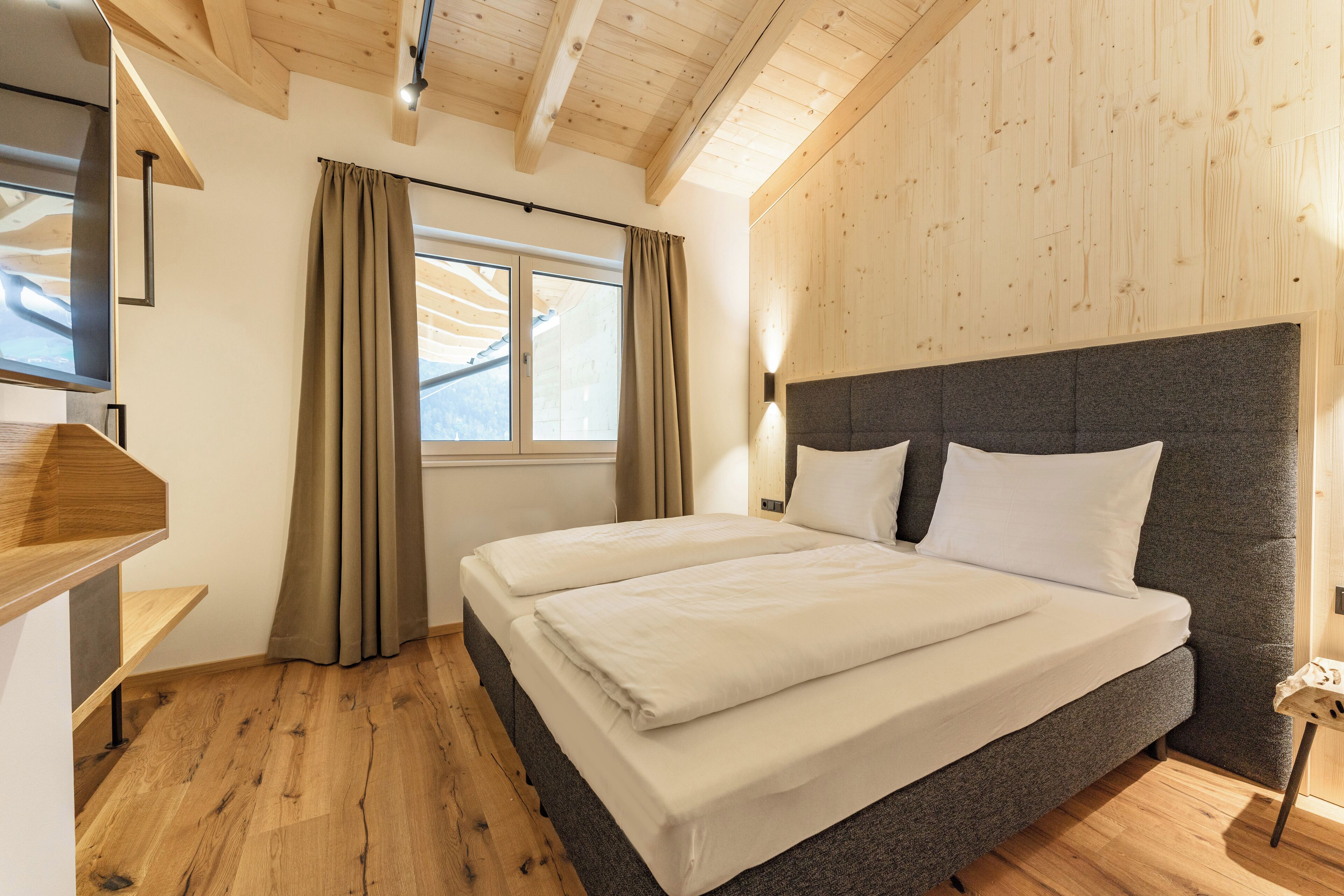 A bedroom with a large bed, wooden flooring, a window with curtains, and a wooden ceiling.