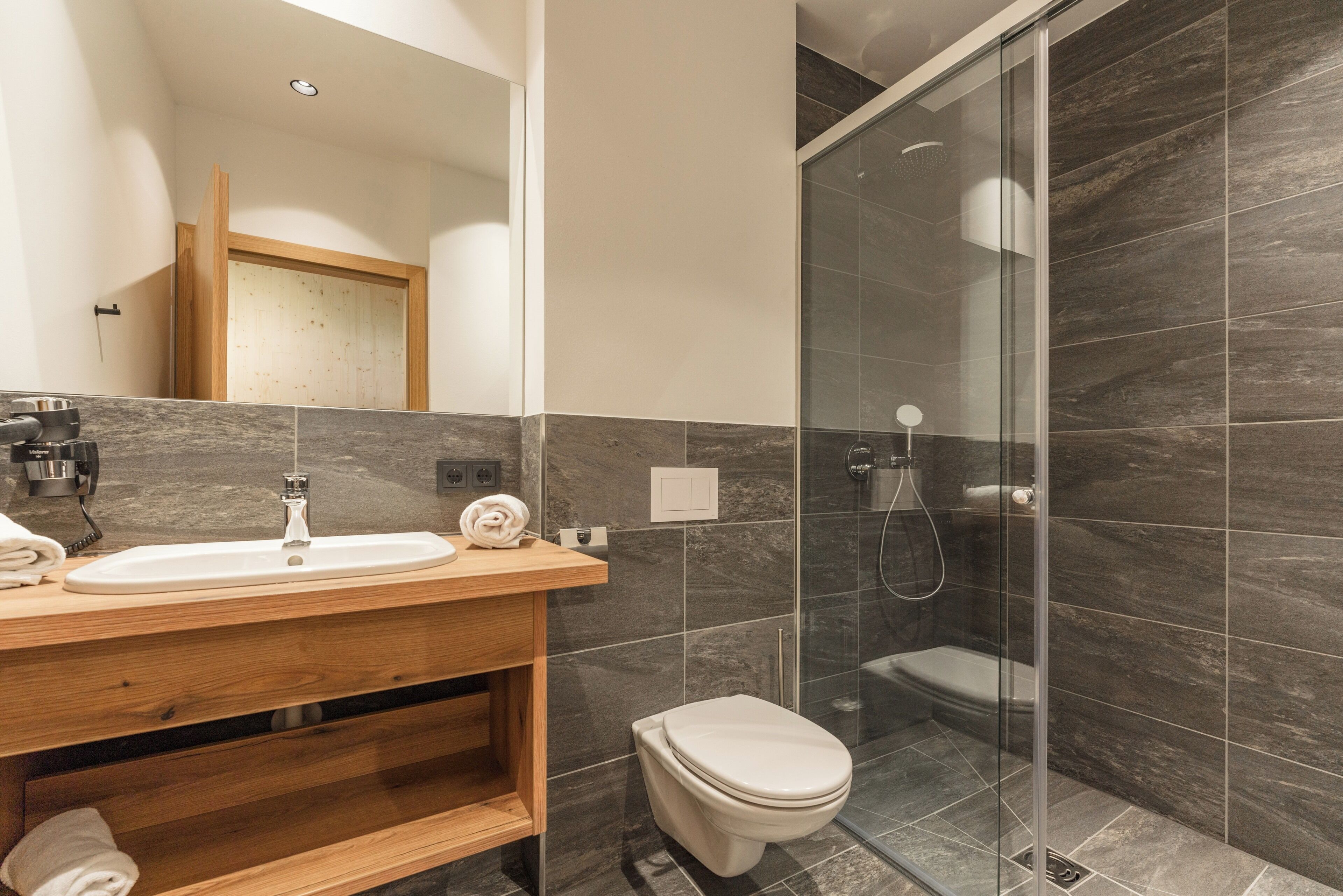 Un baño moderno con tocador de madera, lavamanos blanco, inodoro y zona de ducha con mampara de vidrio.