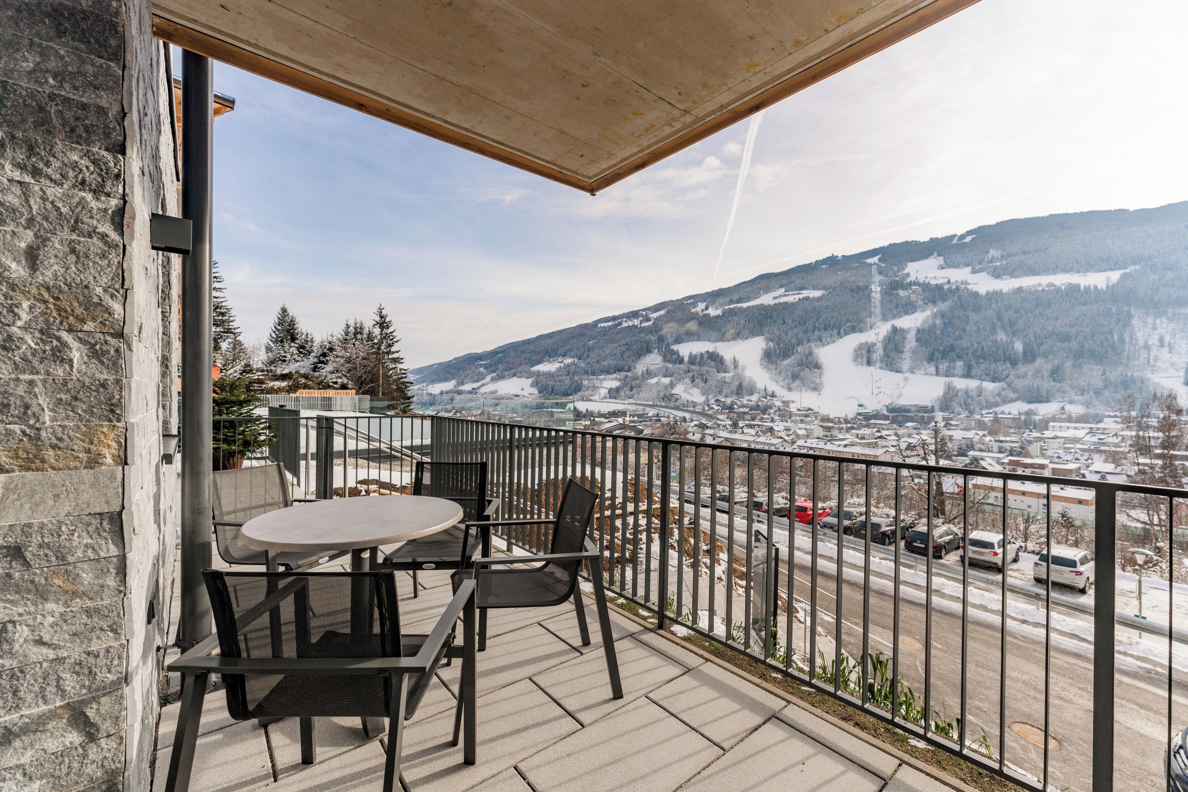 A balcony with a table and chairs, overlooking a snowy mountain and a road with cars.