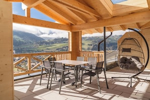 Una terraza de madera con una mesa y sillas, un sillón colgante y vistas a un paisaje montañoso.