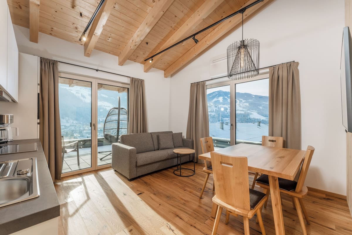 A modern living room with a sofa, dining table, and chairs, large windows with curtains, and a view of mountains.