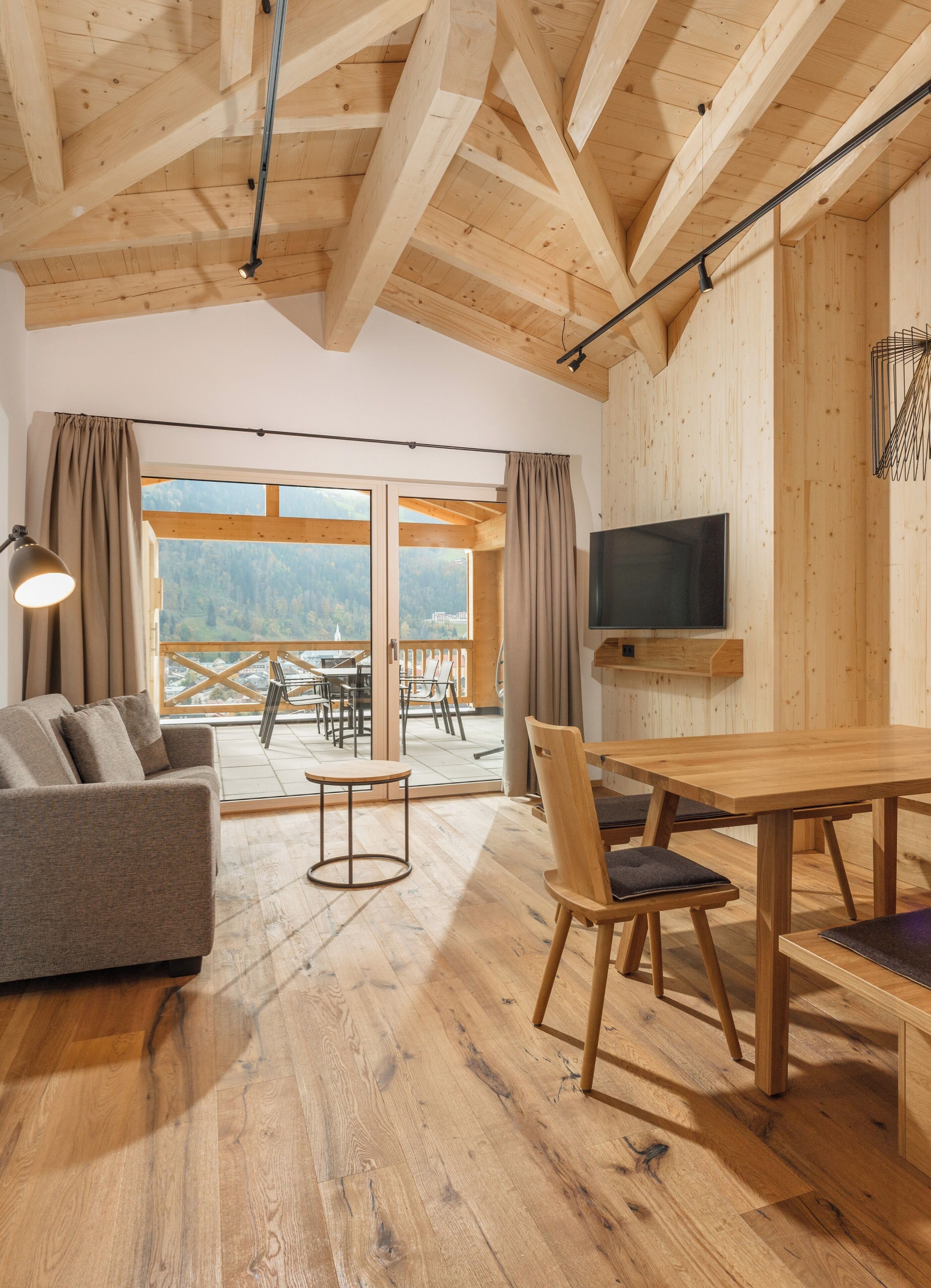 A modern living room with a wooden ceiling, a sofa, a dining table with chairs, a TV mounted on the wall, and large windows offering a view of the outdoors.