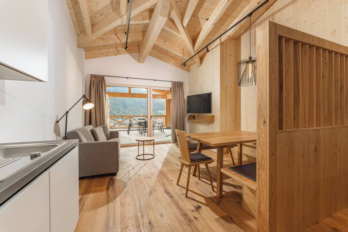 A modern interior with a wooden ceiling, a dining area with a table and chairs, a sofa, a TV mounted on the wall, and a view through large windows.