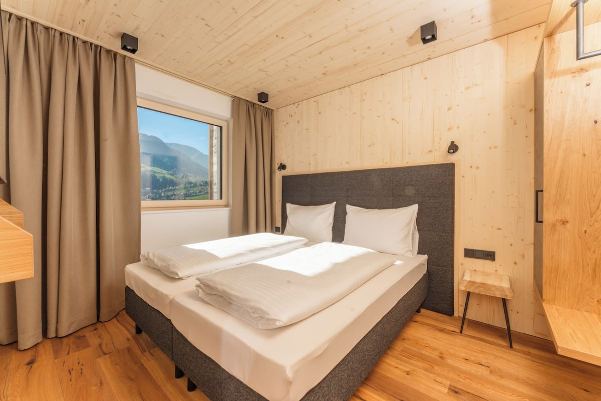 A modern bedroom with a wooden ceiling, a large bed with white linens, a view of mountains through a window, and a wooden wardrobe.