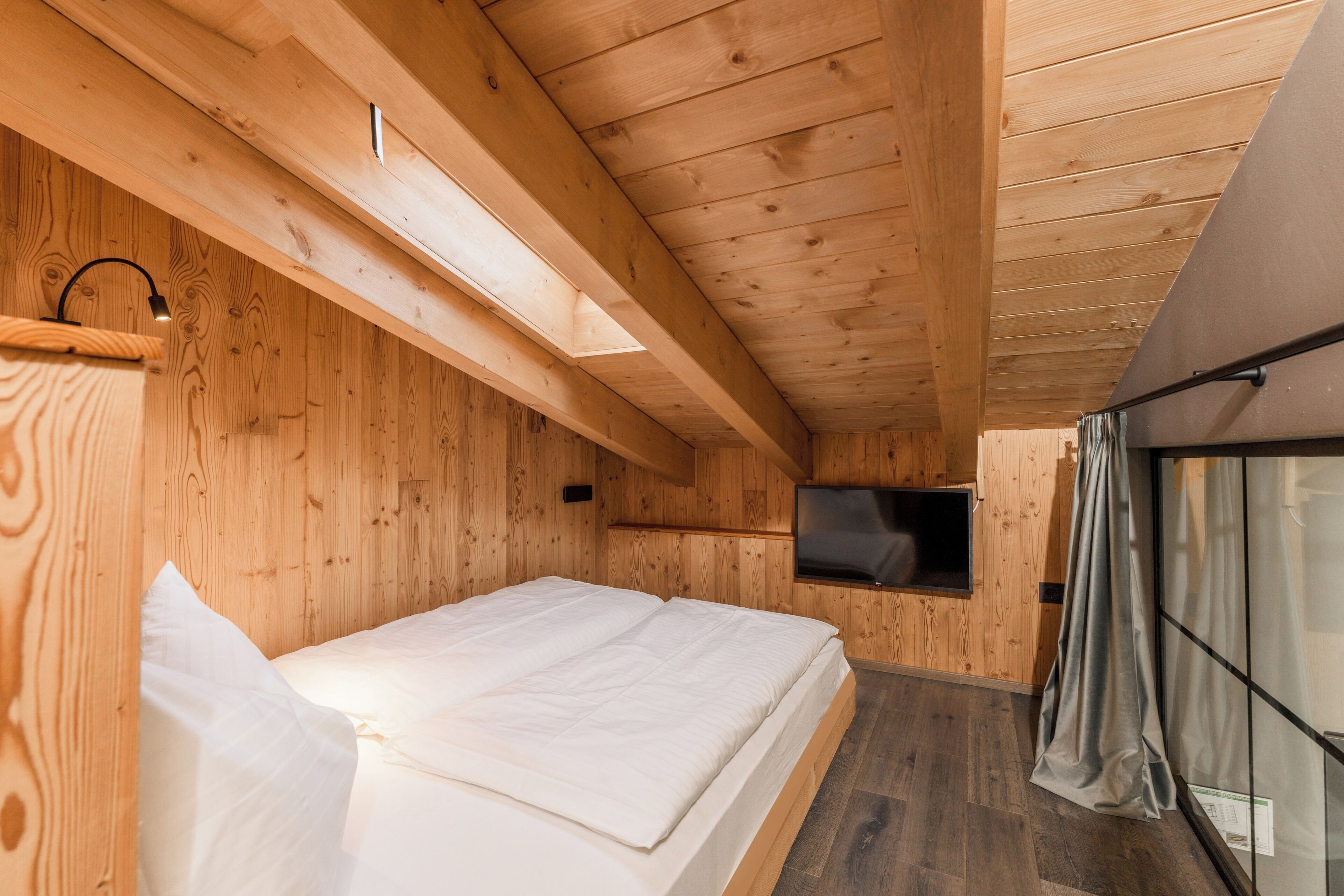 A wooden cabin interior with a bed, a TV mounted on the wall, and a window with curtains.