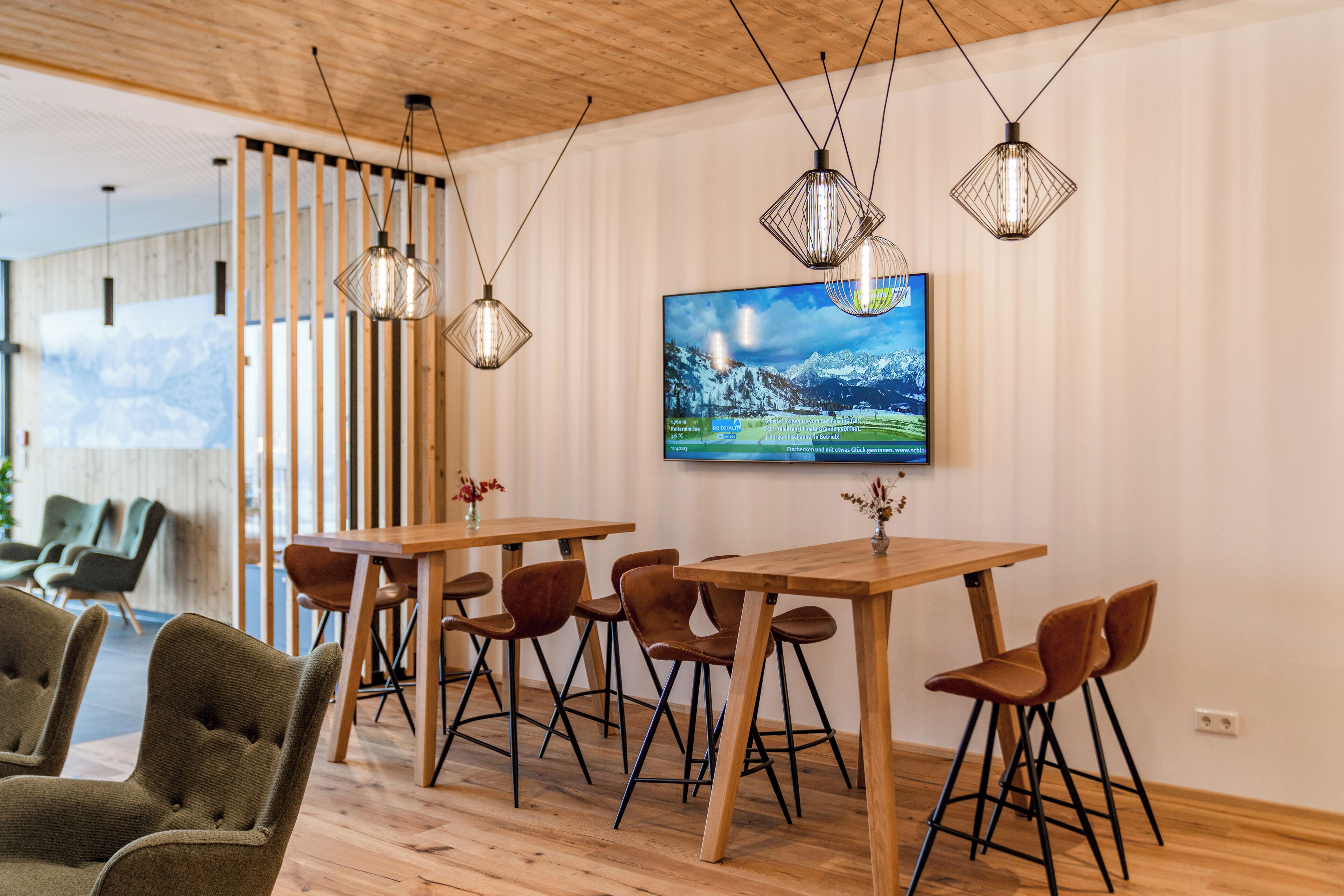 A modern interior with wooden tables and chairs, a large flat-screen TV on the wall, and hanging pendant lights.