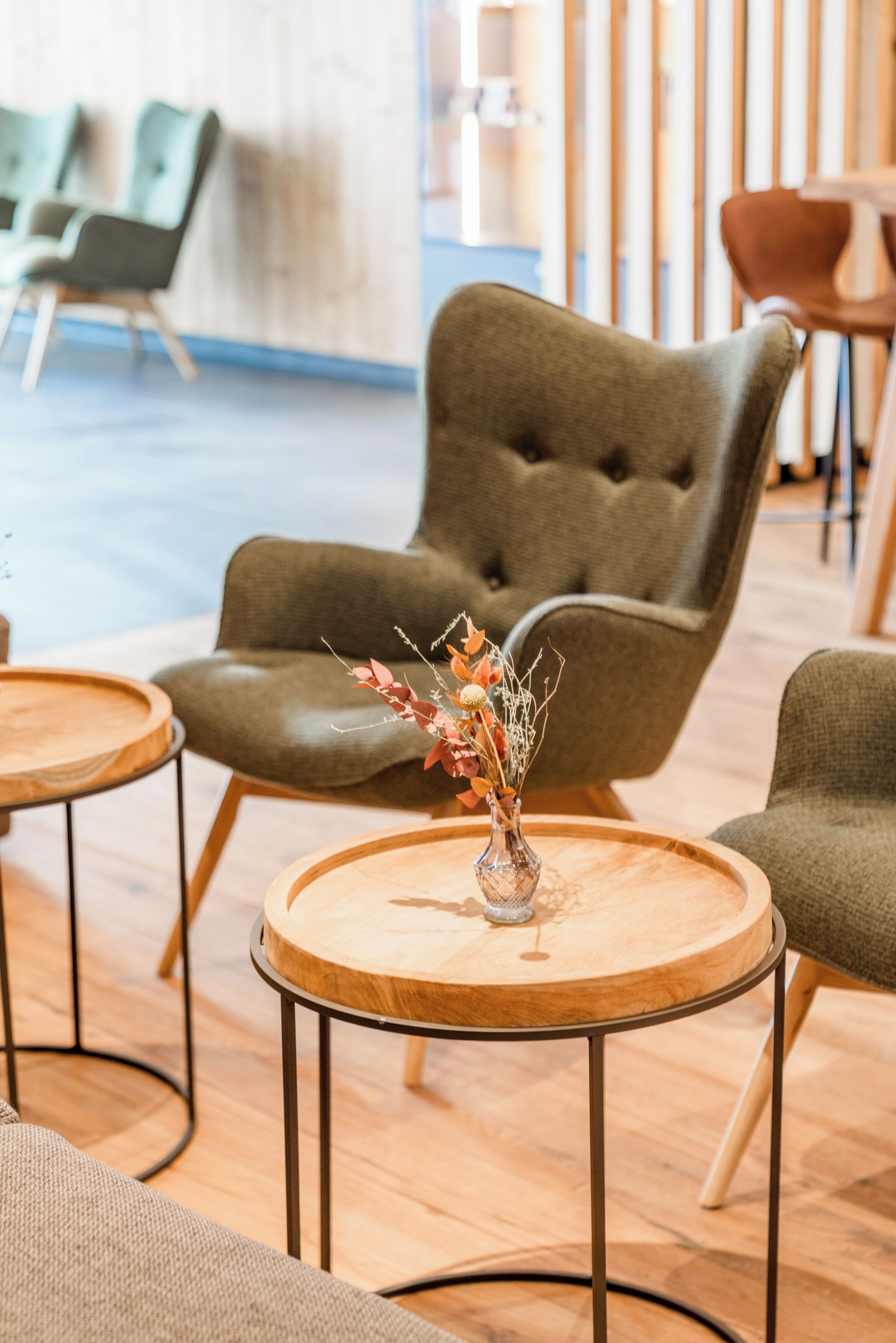 A modern interior with a tufted armchair, round wooden tables, and a vase with dried flowers.