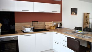 Cocina moderna con armarios blancos, un lavamanos negro y una encimera de madera. Hay una zona de comedor con una mesa y sillas, y un estante con objetos decorativos.