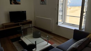 Una sala de estar con televisión, un sofá, una mesita de centro de vidrio y una ventana con vista a la playa.