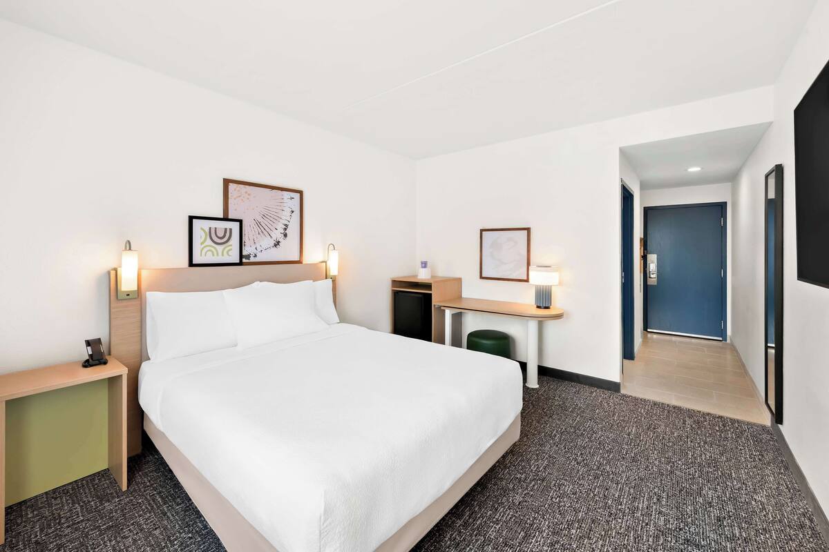 A hotel room with a bed, bedside tables, a desk, and a chair. There are framed pictures on the wall, and a door leading to another room.