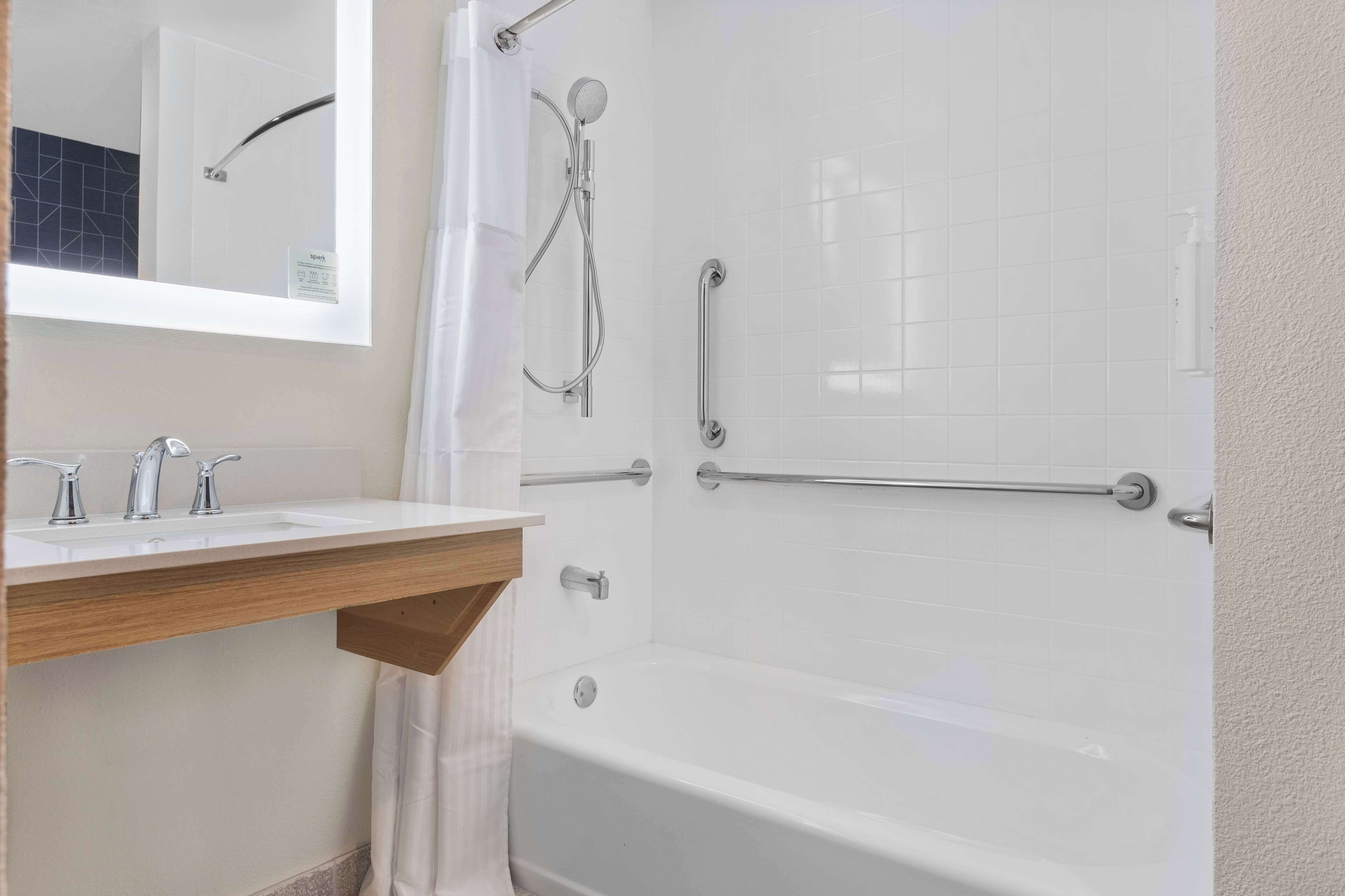 Un baño con un lavabo blanco, un espejo, una ducha con cabezal de ducha de mano y una barra de apoyo.