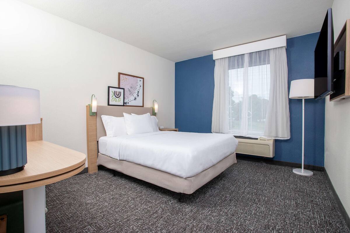 A hotel room with a bed, a desk, a lamp, a TV, and a window with curtains.