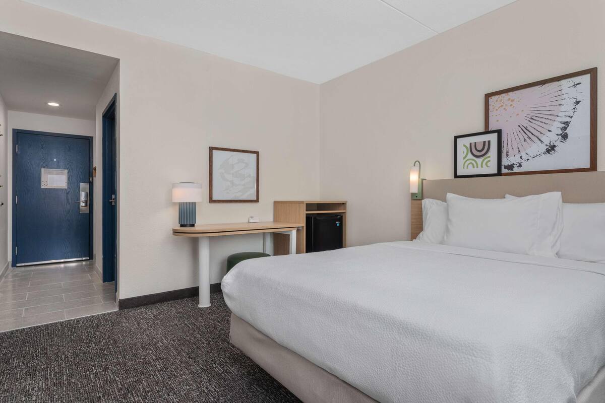 A hotel room with a bed, a desk, a chair, a small table, a wall-mounted lamp, and a door.