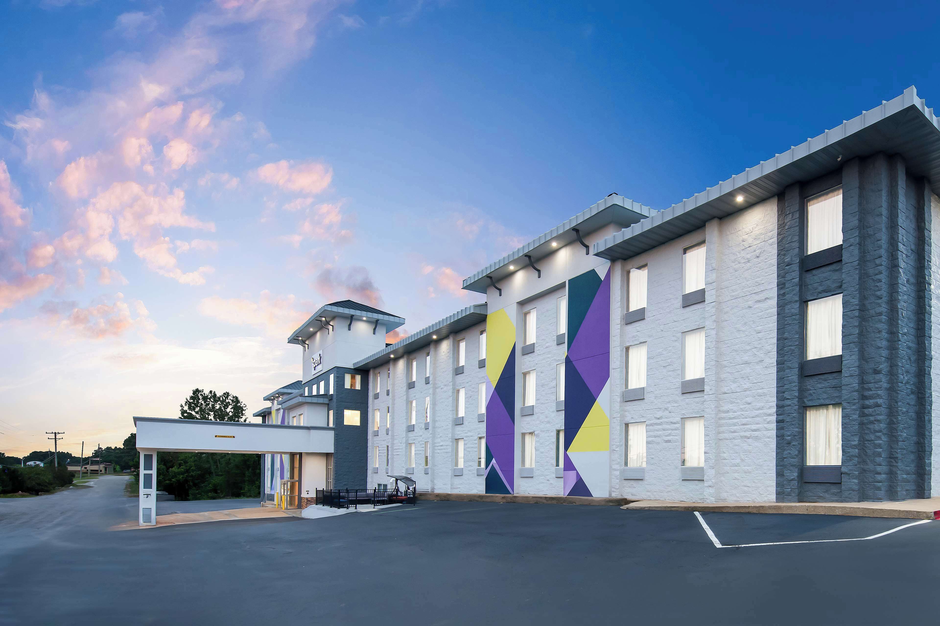Ein modernes Hotel mit großem Parkplatz, überdachter Einfahrt und einer auffälligen geometrischen Wandmalerei an der Fassade.