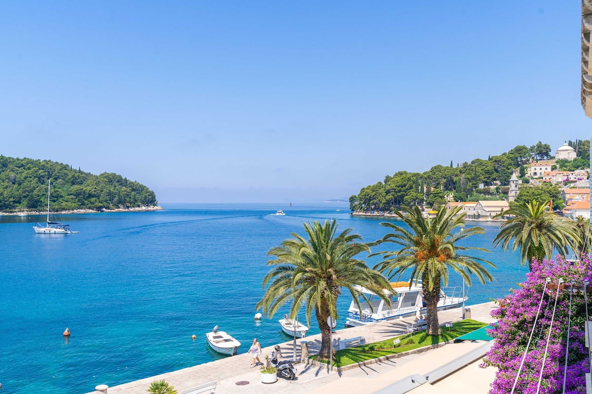 A coastal town with boats docked along the waterfront, palm trees, and a clear blue sea.