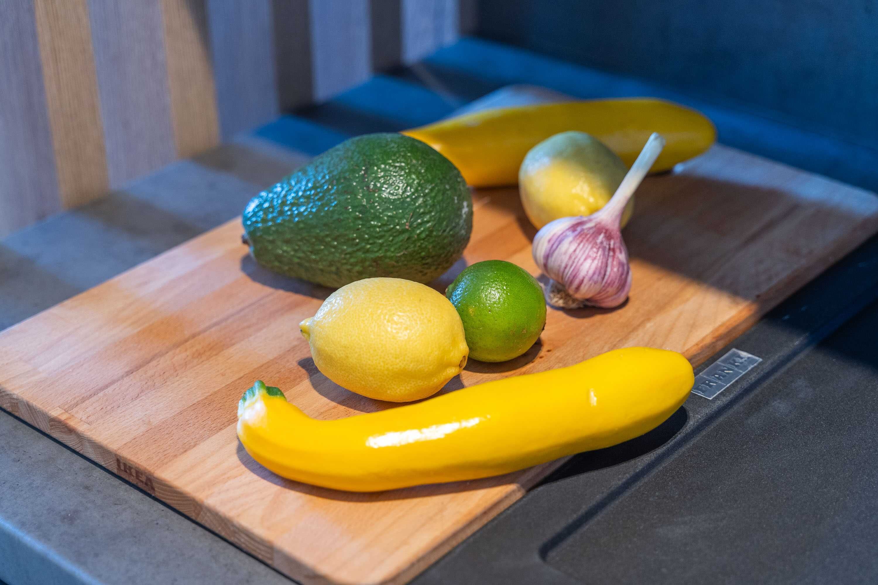 A wooden cutting board with various fruits and vegetables including an avocado, lemons, limes, garlic, and a yellow bell pepper.