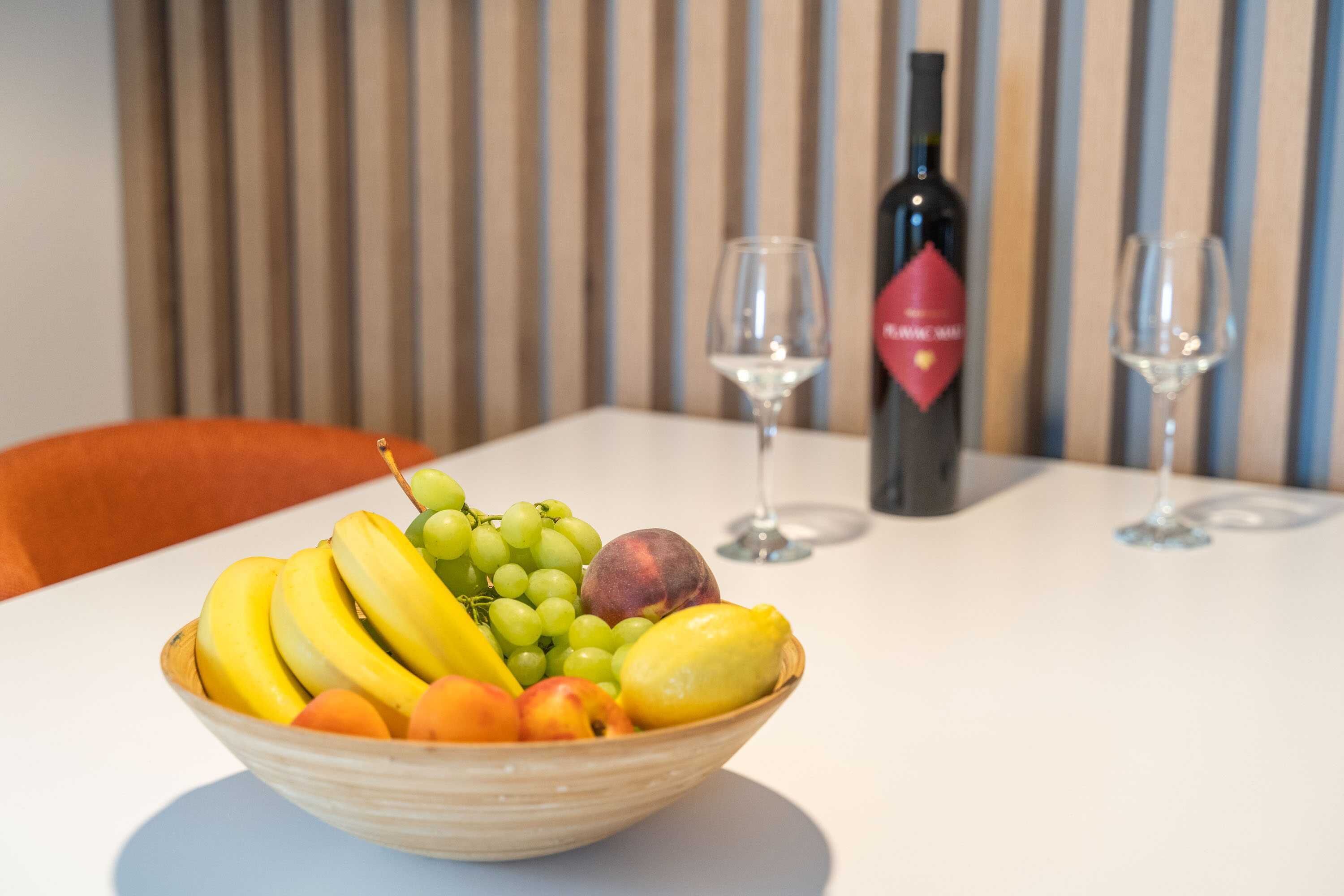 A bowl of fruit on a table with a wine bottle and glasses in the background.