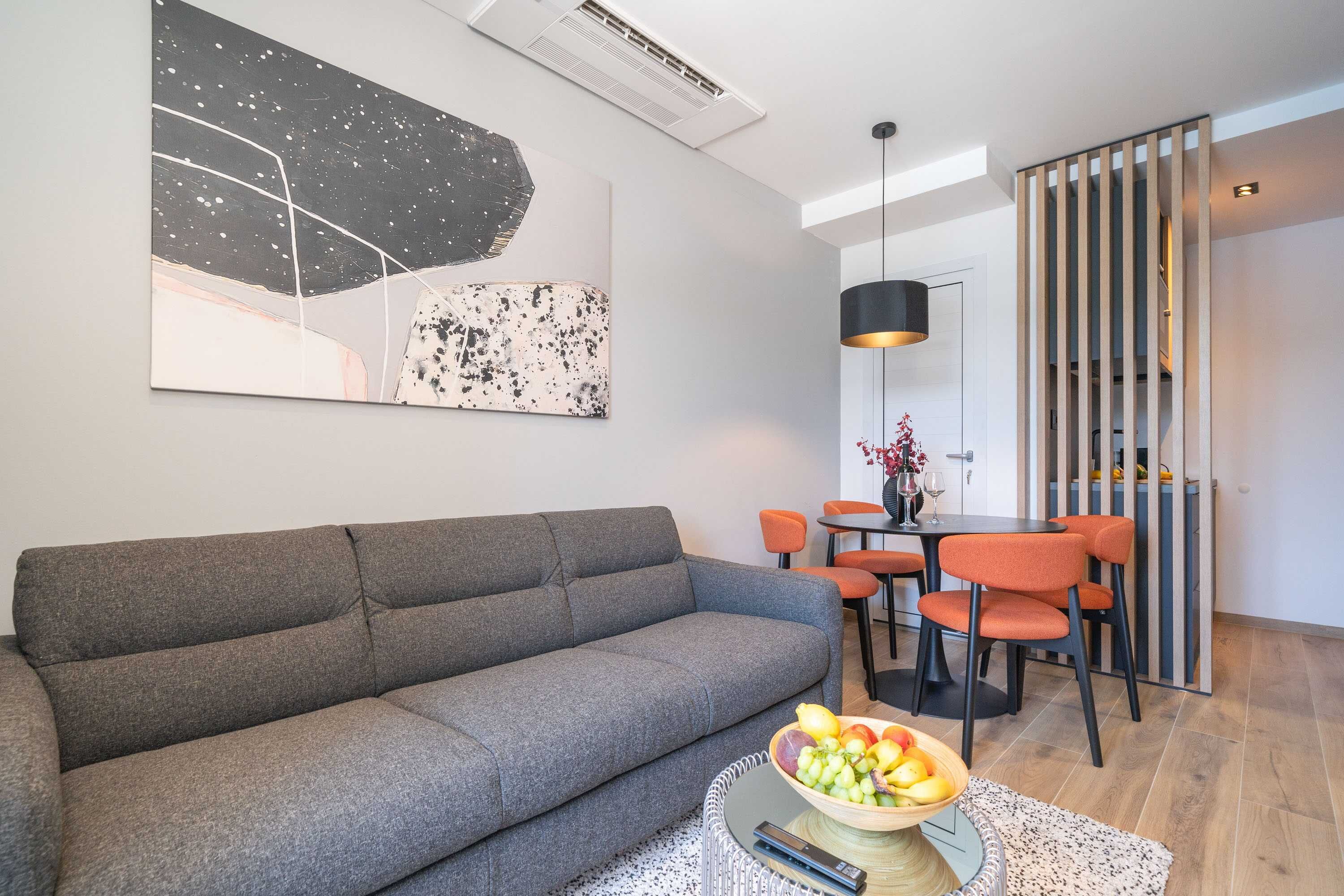 A modern living room with a grey sofa, a dining area with orange chairs, and a fruit bowl on a coffee table.