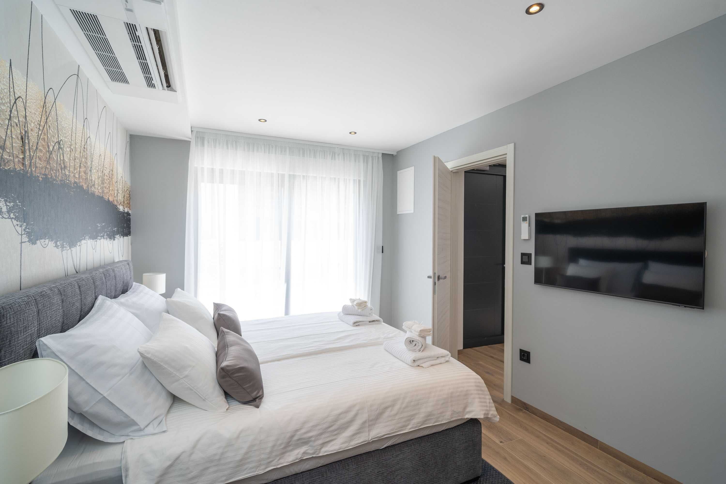 A modern hotel room with a large bed, a flat-screen TV, and a large window with sheer curtains.