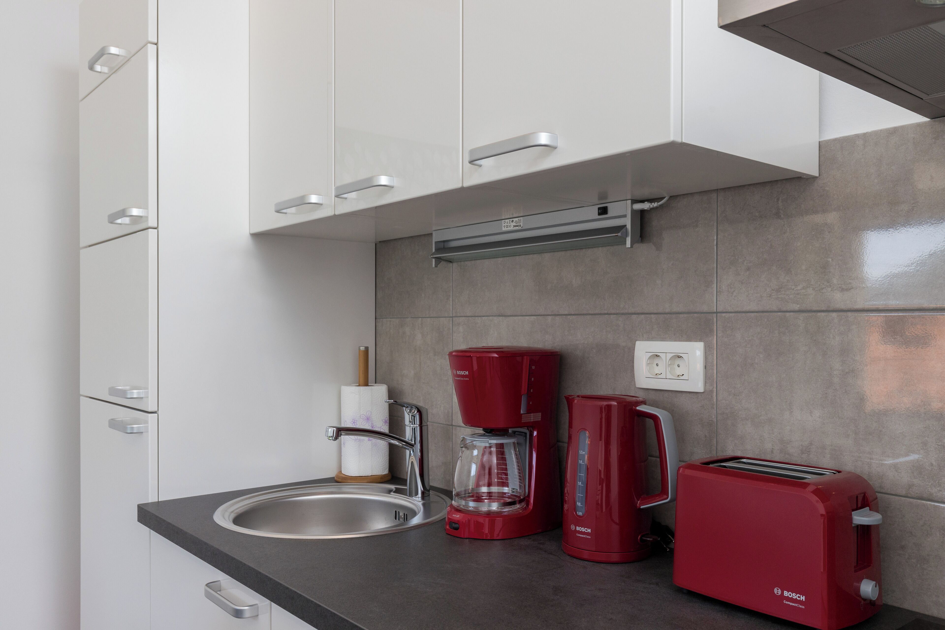 A modern kitchen with white cabinets, a stainless steel sink, a red Bosch coffee maker, a red Bosch toaster, and a glass coffee pot.