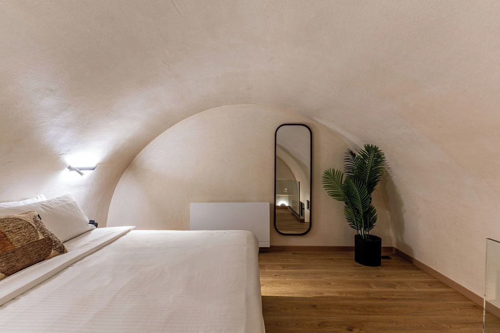 A bedroom with a curved ceiling, a bed with white linens, a mirror, a potted plant, and a wooden floor.