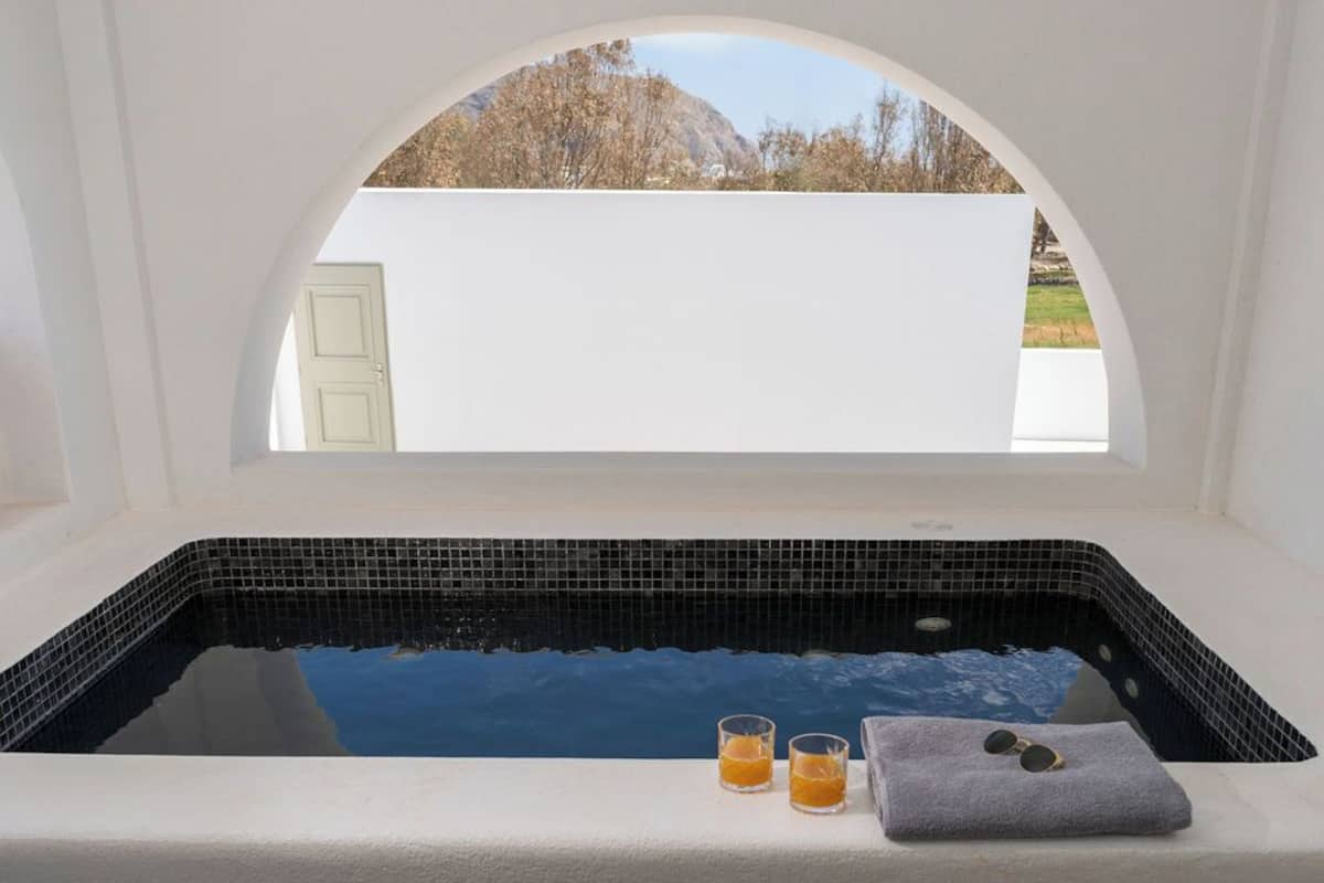 A spa tub with black mosaic tiles, two glasses of orange liquid, and a towel with sunglasses on a white surface.