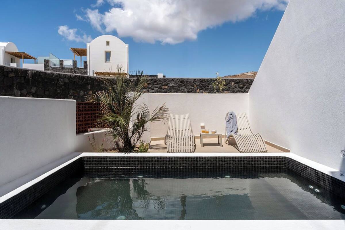 A rooftop pool area with two lounge chairs and a small table, surrounded by white walls and a stone wall.