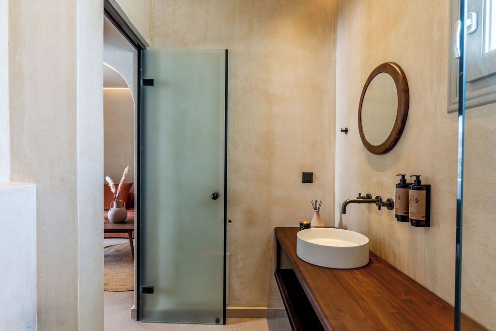 Un baño moderno con puerta de vidrio, espejo redondo y tocador de madera con lavamanos blanco.