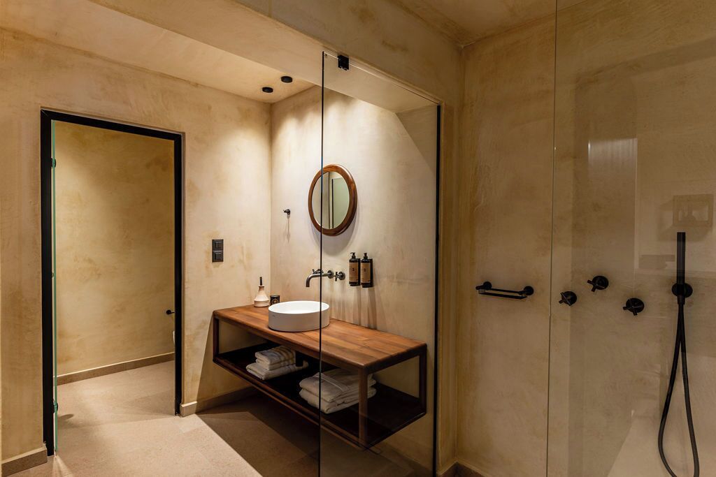 A modern bathroom with a glass shower enclosure, a wooden vanity with a round mirror, and a shelf with folded towels.