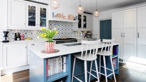 A modern kitchen with white cabinets, a blue island with bar stools, pendant lights, and a patterned backsplash. - Sunlit Symphony, by Veeve (London)