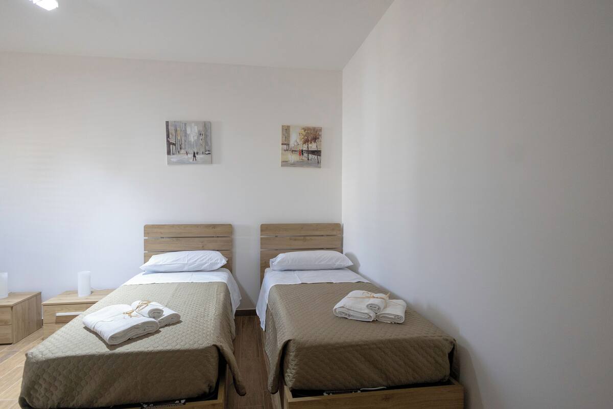 A room with two single beds, each with a wooden headboard, white bedding, and rolled towels. There are two wall-mounted paintings and a small wooden nightstand with a lamp.