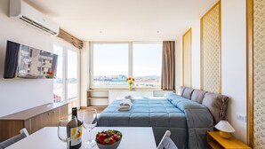 A hotel room with a bed, a dining table with wine glasses and a bowl of fruit, a TV, and a large window with a city view. - Chiave di Fuga - Doppia Deluxe (Naples)