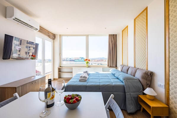 Quarto de hotel com cama, mesa de jantar com copos de vinho e uma tigela de frutas, televisão e uma janela grande com vista para a cidade.