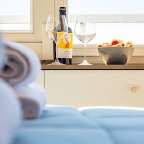 A neatly made bed with a white pillow and a rolled towel. A bottle of wine, a wine glass, and a bowl of fruit are placed on a wooden surface near a window. - Chiave di Fuga - Doppia Superior (Naples)