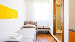 Una habitación pequeña y luminosa con una cama, una mesita de noche con lámpara y una zona de ducha visible a través de un vidrio.