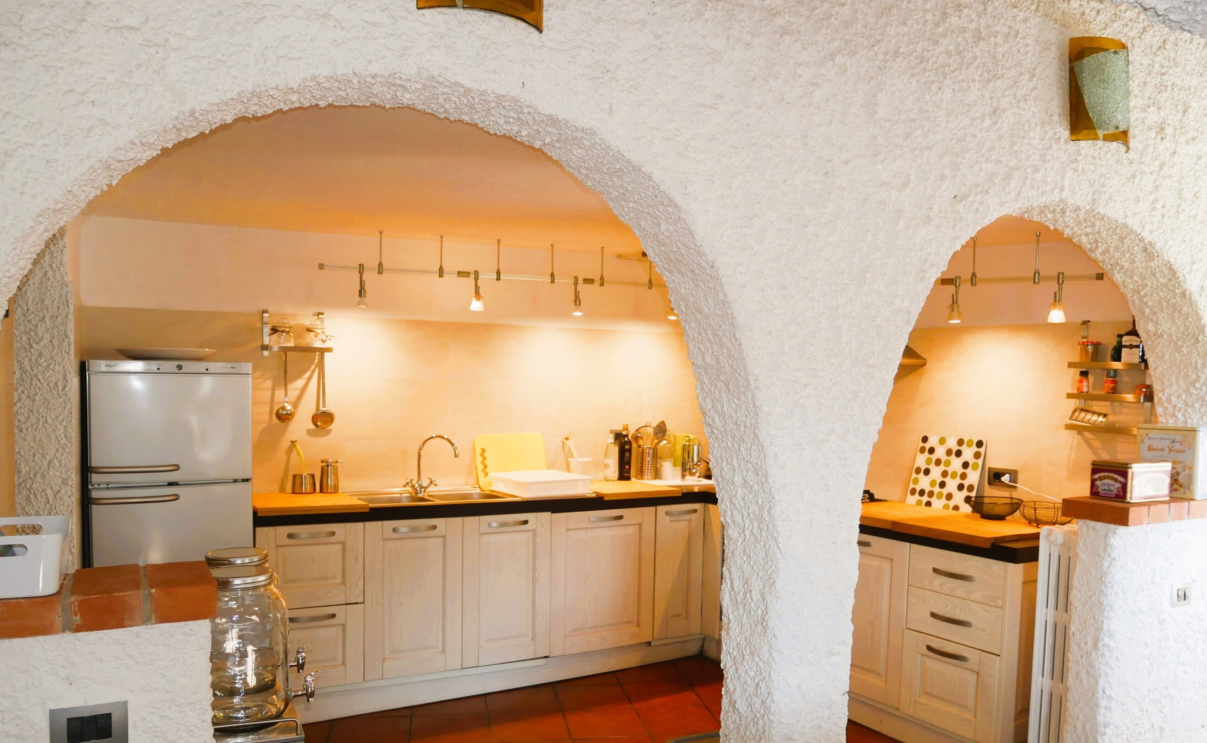 A kitchen with white cabinets, a double sink, and a refrigerator. There are hanging lights and a shelf with kitchen items.
